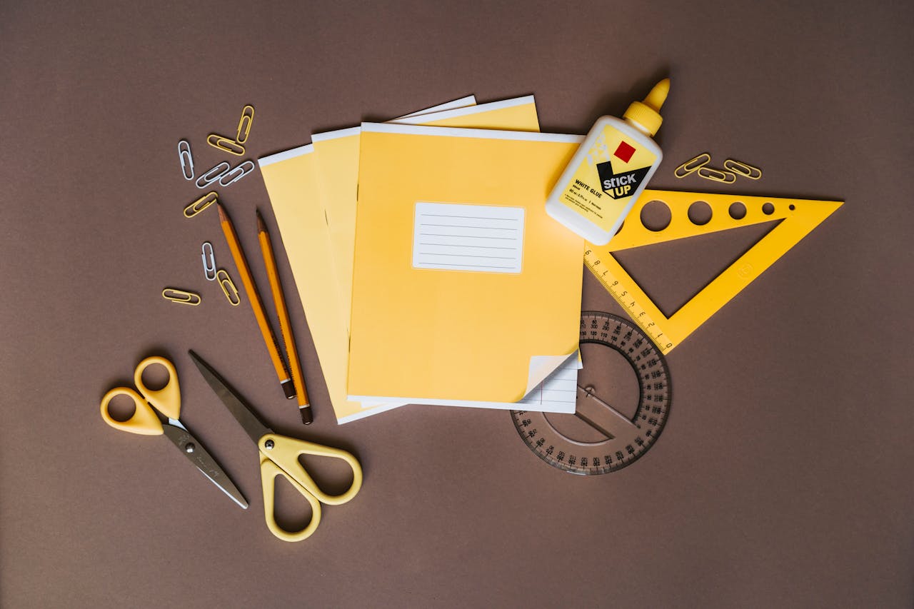 A flat lay of yellow stationery tools like pencils, scissors, and glue on a brown surface.